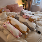 Snug Cat long plushies in every size and colour stacked on a bed